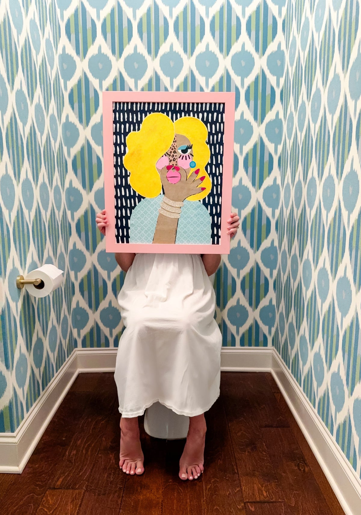 A person in a white dress sits barefoot on a toilet in a bathroom with blue and white patterned wallpaper, holding a framed pop-art portrait of a woman with bright yellow hair and plugging her nose.